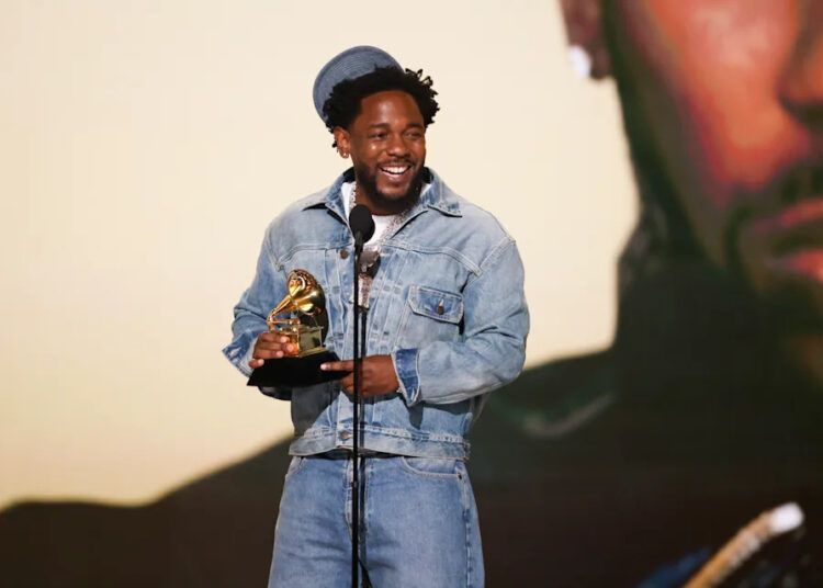 Kendrick Lamar in denim holds a Grammy award, smiling on stage. He could win the best album of the year grammy for gnx.