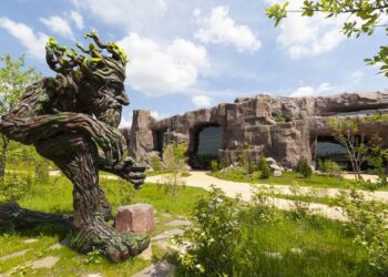 Large tree-like sculpture in a garden with a rocky building background under a blue sky.
