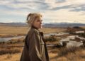 A woman in an oversized jacket stands in a field with a flowing creek seen in the background.