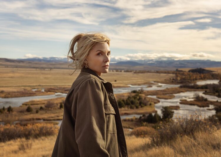 A woman in an oversized jacket stands in a field with a flowing creek seen in the background.