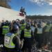 White truck with two people sat on the roof - dozens of police officers in riot gear are standing in a group with a large crown seen beyond in a field.