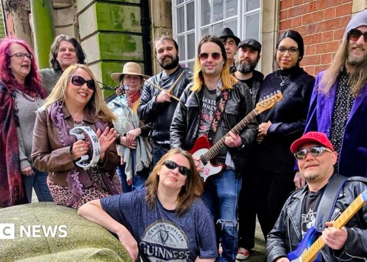 A group of 12 musicians stand with musical instruments including guitars and a tambourine on a street in Walsall, smiling at the camera.