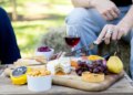 A chopping board filled with cheeses, grapes, a pear and more, a glass of wine behind, and a person's hand holding a cheese knife about to cut into a cheese.