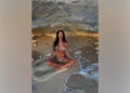 Nicole Scherzinger poses in a zebra bikini while meditating on the beach under a rock formation with her legs crossed.
