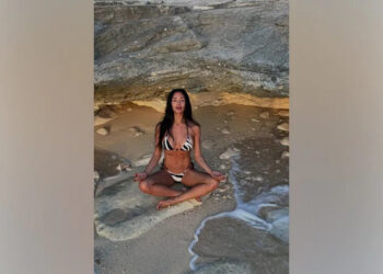 Nicole Scherzinger poses in a zebra bikini while meditating on the beach under a rock formation with her legs crossed.