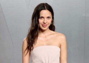 A photograph of Olivia Rodrigo posing for a picture in a plain, cream colored strapless dress before a gray backgroumd