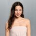 A photograph of Olivia Rodrigo posing for a picture in a plain, cream colored strapless dress before a gray backgroumd