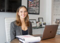 A woman at a conference room table with a laptop computer and legal pad. She is smiling at the camera.