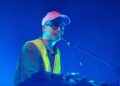 Alexis Taylor of Hot Chip performs on stage, wearing a pink cap, high vis jacket and shirt, while playing the keyboard and singing into a microphone