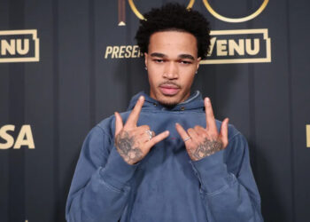 PlaqueBoyMax in a blue hoodie poses with hand gestures in front of a backdrop with logos like VENU, Visa, and Billboard.