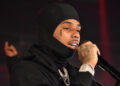 Pooh Shiesty wearing a black hoodie and balaclava, holding a microphone. He has a teardrop tattoo and visible jewelry.