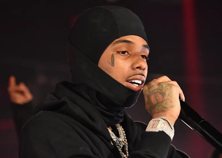 Pooh Shiesty wearing a black hoodie and balaclava, holding a microphone. He has a teardrop tattoo and visible jewelry.