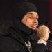 Pooh Shiesty wearing a black hoodie and balaclava, holding a microphone. He has a teardrop tattoo and visible jewelry.