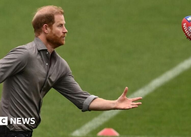 Prince Harry is pictured in a dark suit reaching out to place a poppy on a memorial wall commemorating Australian war dead from the Afghanistan conflict