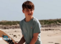 A grinning Louis is shown leaning with his arms folded against a metal barrier, with the sea as the backdrop.