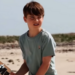 A grinning Louis is shown leaning with his arms folded against a metal barrier, with the sea as the backdrop.