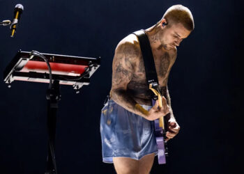 Justin Bieber plays a guitar on stage, wearing shorts, with a keyboard nearby.