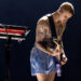 Justin Bieber plays a guitar on stage, wearing shorts, with a keyboard nearby.