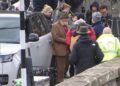 Sir Anthony Hopkins is stood by a Range Rover wearing a brown suit jacket and trousers, and a hat. He is surrounded by people from a production company for his new film.