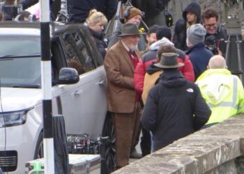 Sir Anthony Hopkins is stood by a Range Rover wearing a brown suit jacket and trousers, and a hat. He is surrounded by people from a production company for his new film.
