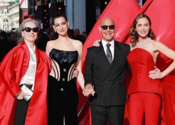 Meryl Streep, Anne Hathaway, Stanley Tucci and Emily Blunt pose on the red carpet in London. Streep is wearing sunglasses, a long, red, satin jacket, a cream blouse and black trousers. Hathaway has her hair up and is wearing a black embellished corset. Tucci is wearing a pinstriped black suit, white shirt and tie, along with sunglasses. Blunt has a red two-piece on an a jewelled necklace.