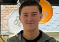 An 18-year-old man standing in front of orange, white and black balloons by a window. He has short dark hair and is smiling. Wearing a blue jacket and a black top underneath.