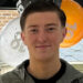 An 18-year-old man standing in front of orange, white and black balloons by a window. He has short dark hair and is smiling. Wearing a blue jacket and a black top underneath.