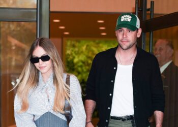 Two individuals, one dressed in a white shirt and black jacket, the other in a light-colored long-sleeved top and sunglasses, are standing side by side and appear to be leaving a building.