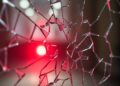 An abstract close-up photograph of shattered glass reflecting a faint red light, conveying a sense of disruption and exposure surrounding the political elite.