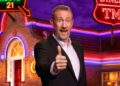 Alex Horne wearing a suit jacket and shirt, giving a thumbs-up sign and big smile, standing outside the Taskmaster house.