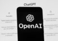 The OpenAI logo is seen on a phone in front of a computer screen displaying ChatGPT.