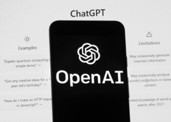 The OpenAI logo is seen on a phone in front of a computer screen displaying ChatGPT.