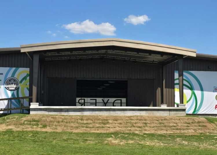An outdoor stage with a brown roof and banners displaying various logos stands on a grassy area under a blue sky. "DYER" is visible on the stage floor, and a bird is flying in the upper right corner of the image.