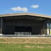 An outdoor stage with a brown roof and banners displaying various logos stands on a grassy area under a blue sky. "DYER" is visible on the stage floor, and a bird is flying in the upper right corner of the image.