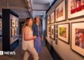 Two women with blonde hair in an art gallery looking at photographs of the British royal family on a wall.