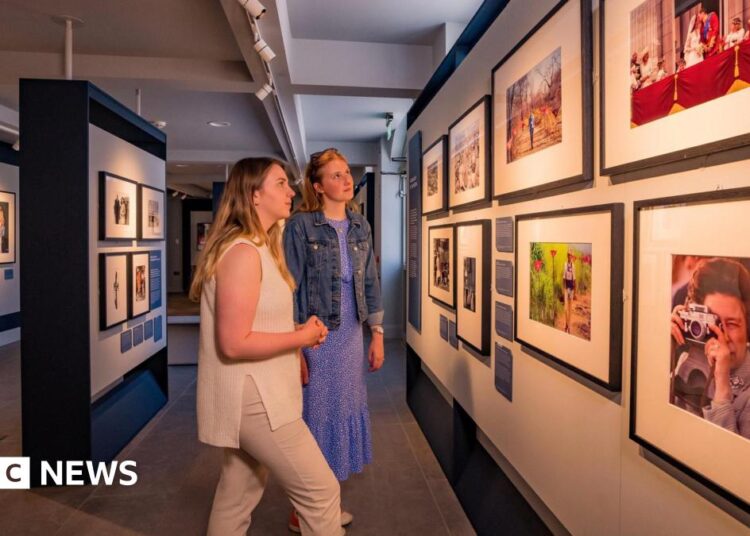 Two women with blonde hair in an art gallery looking at photographs of the British royal family on a wall.