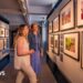 Two women with blonde hair in an art gallery looking at photographs of the British royal family on a wall.