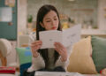 A woman (Jisoo of Blackpink) reads a Korean device manual while sitting on a couch in an apartment, in the Netflix K-drama 'Boyfriend on Demand.'