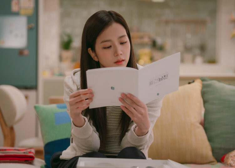 A woman (Jisoo of Blackpink) reads a Korean device manual while sitting on a couch in an apartment, in the Netflix K-drama 'Boyfriend on Demand.'