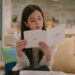 A woman (Jisoo of Blackpink) reads a Korean device manual while sitting on a couch in an apartment, in the Netflix K-drama 'Boyfriend on Demand.'