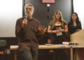 A man in a brown shirt speaks in front of a white and black wall and two women.