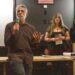 A man in a brown shirt speaks in front of a white and black wall and two women.