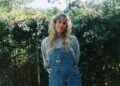 A woman wearing overalls with long curly blonde hair smiles among foliage.