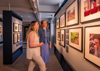 Two women with blonde hair in an art gallery looking at photographs of the British royal family on a wall.