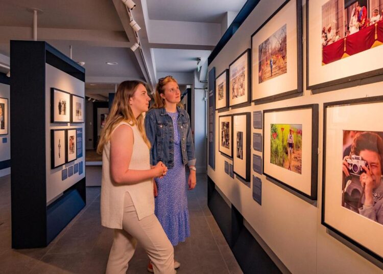 Two women with blonde hair in an art gallery looking at photographs of the British royal family on a wall.