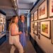 Two women with blonde hair in an art gallery looking at photographs of the British royal family on a wall.