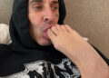 Travis Barker is lying down, wearing a black hoodie, playfully holding a foot to his mouth.