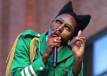 Tyler, The Creator in a green outfit with cat-ear hairstyle and mask gestures while holding a microphone.