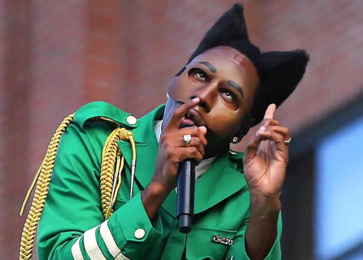 Tyler, The Creator in a green outfit with cat-ear hairstyle and mask gestures while holding a microphone.