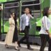 Prince Harry and Meghan Markle disembark a tram in Melbourne on a royal visit to Australia. Harry, a tall man with red hair and beard, is dressed in a grey blazer with black slacks. Megan, a woman with dark hair, is wearing a black dress with a tan coloured trenchcoat and ballet flats.